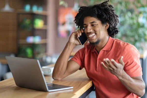 Uzaktan kumanda. Cafe 'de dizüstü bilgisayar kullanan ve cep telefonuyla konuşan neşeli siyahi genç adam, Milenyum Afrikalı Amerikalı Serbest Çalışan ve Cep Telefonu Konuşmasının tadını çıkaran,
