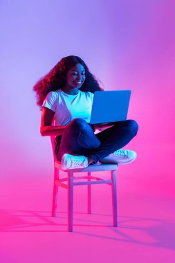 Mutlu genç siyahi kadın bir sandalyede otururken çevrimiçi çalışıyor ya da ders çalışıyor. Neon ışığı, uzak bir video konferansına katıldığı için canlı bir atmosfer yaratıyor..