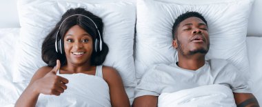 Bir erkek yatakta huzur içinde uyurken, kulaklık takan bir kadın başparmağını kaldırıyor. Enerjik ve mutlu görünüyor. Sahne yumuşak yastıklarla dolu..