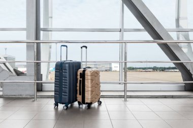 Havalimanında panoramik pencerenin yanında duran iki şık plastik bavul, Terminalde bekleyen mavi ve bej seyahat çantaları, Ulaşım ve Seyahat Konsepti için yaratıcı çekim,