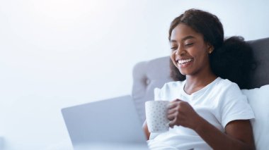 Kanepede oturan bir kadın elinde gülümseyerek bir fincan çay tutuyor. Önündeki dizüstü bilgisayara odaklanmış. Odanın aydınlığı ve yumuşak bir atmosferi var..