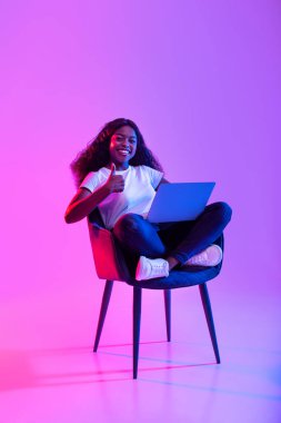 Genç bir Afrikalı Amerikalı kadın bir koltukta oturuyor ve dizüstü bilgisayarı üzerinde çalışıyor. Olumlu bir şekilde gülümsüyor ve parlak bir neon arka plana başparmak işareti yapıyor, uzak çalışmadaki neşesini vurguluyor..