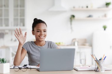 Gülümseyen Afro-Amerikan liseli kız bilgisayarda öğretmenine sesleniyor ve el sallayarak minimalist mutfak içinde web kamerasına el sallıyor. Sosyal mesafe, uzak ders, toplantı, covid-19 salgını