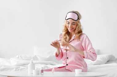 Pembe pijamalı, gülümseyen Avrupalı sarışın kadın. Maskeli, tırnaklarını boyuyor. Yatak odasının iç kısmında oturuyor, fotokopi çekiyor. Pozitif duygular, ev güzellik bakımı ve covid-19 karantina