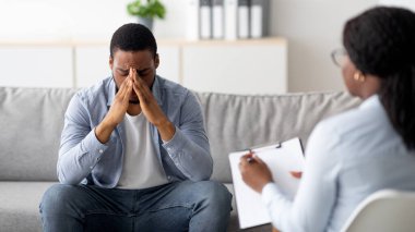 Umutsuz bir siyah adam ruhsal çöküntü ya da depresyondan muzdarip. Psikolog ofisinde, panoramada. Stresli erkek hasta terapistle görüşüyor, profesyonel yardım alıyor.