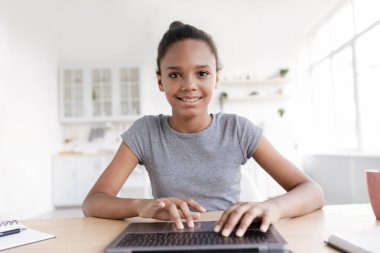 Modern öğrenci ve eğitim için aletler. Ev çalışması, sohbet ve mesafeli konuşma. Gülümseyen Afro-Amerikalı kız kameraya bakıyor ve oturma odasında dizüstü bilgisayarla klavyeye yazı yazıyor.