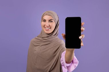 Kafiyeli mutlu Müslüman kadının ellerinde siyah boş ekranlı akıllı telefonun görüntüsü, menekşe arkaplan üzerinden kameraya gülümsüyor, seçici odaklanma