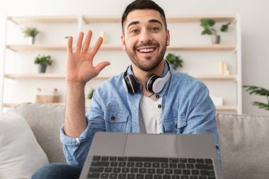 Çevrimiçi Video Çağrısı. Gülümseyen sakallı bir adamın portresi arkadaşlarıyla, ailesiyle ya da meslektaşlarıyla bilgisayar kullanarak sanal görüşme yapıyor, el sallıyor, evdeki koltukta oturuyor, web kamerası görüntüsü var.