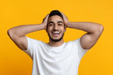 Süper mutlu, orta doğulu beyaz tişörtlü, kameraya gülümseyen ve kafasına dokunan yakışıklı adam, gerçekten iyi haberler aldı, kendinden geçmiş, sarı stüdyo arka planı, yakın portre