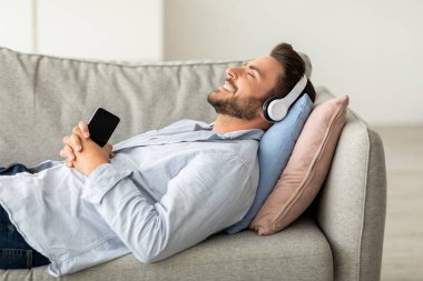 Kablosuz kulaklıklı mutlu adam kanepeye uzanıyor, gözleri kapalı ve gülümsüyor, evde müzik dinlerken, akıllı telefon tutuyor, yeni stereo kulaklıkların keyfini çıkarıyor.