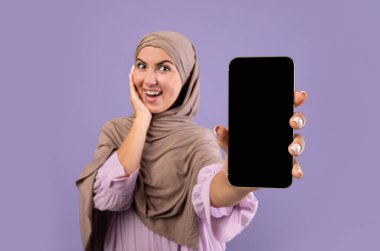 Siyah ekranlı akıllı telefonun görüntüsü tesettürlü mutlu Müslüman kadının ellerinde, mor arkaplan üzerinde poz veriyor, seçici odak, boş alan
