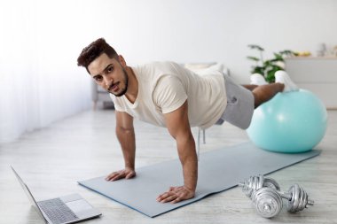 Çekici genç Arap çocuk dizüstü bilgisayarın yanında duruyor, şınav çekmek için fitness topunu kullanıyor, online spor eğitmenliği yapıyor, evde karın kaslarını çalıştırıyor. Yurtiçi eğitim kavramı