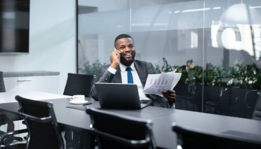 Modern bir ofiste konferans masasında oturan bir iş adamı. Telefonda konuşuyor ve elinde belgeler var. Yanına bir laptop ve bir fincan konur..