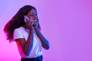 Çekici genç siyahi bir kadın canlı neon ışıkları altında bir telefon konuşmasında mutlu bir şekilde yer alıyor. Boş bir alanda coşkuyla iletişim kurarken gülümsüyor..