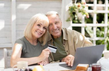 İnternetten alışveriş konsepti. Modern yaşlı çift dizüstü bilgisayar ve kredi kartı kullanıyor, online alışveriş yapıyor, kırsal alanda oturuyor. Heyecanlı müşteriler, e-ticaret konsepti