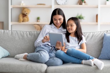 Mutlu Asyalı anne ve kız dijital tablet kullanıyor, video izliyor ya da internette sörf yapıyor, evdeki kanepede oturuyor, kopya çekiyor. Genç Koreli aile birlikte ped kullanıyor.