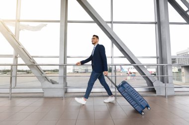 Havaalanı terminalinde pencerenin yanında yürüyen gülümseyen Arap bir iş adamının portresi, bavullarını uçuş kapısına taşıyan yakışıklı bir Orta Doğulu, iş gezisine hazır, kenar manzaralı.