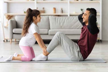 Evde karın kasları üzerinde birlikte çalışan tatlı baba ve kız, babasının bacaklarını tutan sevimli küçük kız, o mekik çekerken, oturma odasında birlikte egzersiz, yan görüş, kopyalama alanı