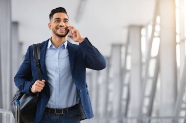 Havaalanı terminalinde yürürken cep telefonuyla konuşan gülümseyen Orta Doğulu adam, mutlu Arap iş adamı el bagajı taşıyor ve cep telefonuyla keyifli sohbetler yapıyor.