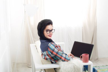Dizüstü bilgisayar ile beyaz koltuk üzerinde koruyucu bilgisayar gözlüklü genç kız. Hipster serbest meslek sahibi kadın evde çalışıyor. Ev ofis, serbest. Beyaz kanepe, modern iç içinde laptop ile esmer kız.