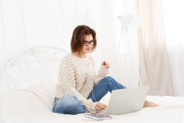 Genç kadın yatakta dizüstü bilgisayar okur. Dalmış kız gözlük ve kulaklık ile hareket eden telefon. Çekici kadın beyaz yatak, rahat kıyafetler içinde. Internet Serbest meslek. Yüksek anahtar, yumuşak sesi