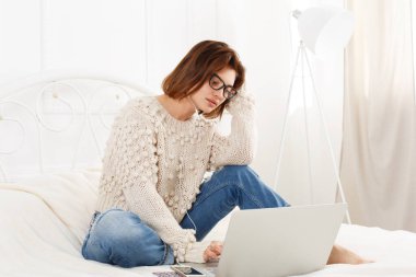 Genç kadın yatakta dizüstü bilgisayar okur. Dalmış kız gözlük ve kulaklık ile hareket eden telefon. Çekici kadın beyaz yatak, rahat kıyafetler içinde. Internet Serbest meslek. Yüksek anahtar, yumuşak sesi