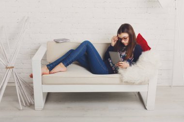 Genç kız eve kanepe tablet bilgisayar ile oturup rahatlayın. Çocuk gözlük, gözlük takıyor. Kız koltukta yalan gadget tablet ile Eyewear. Ergenlik, internet bağımlılığı. Yüksek anahtar