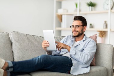 Teknoloji konsepti. Gülümseyen milenyum adamı modern dairesindeki kanepesinde dinlenirken dijital tablet kullanıyor. Mutlu adam internette geziniyor, oturma odasındaki kanepede dinleniyor.