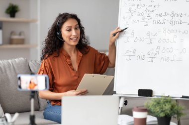 Covid-19 salgını sırasında internetten çalışma. Gülümseyen, kulaklıklı kadın matematik öğretmeni akıllı telefon ve bilgisayar kullanarak internet üzerinden ders veriyor. Vlog kaydeden öğrencilerle video görüşmesi yapan Milenyum Öğretim Üyesi