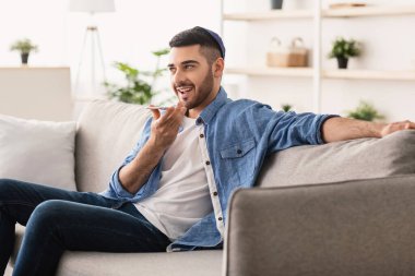 Dijital Hoparlör Uygulaması. Evdeki kanepede oturan genç Yahudi adamın portresi, cep telefonuyla konuşan, sanal ses asistanını kullanarak çevrimiçi arama yapmak ya da hatırlatma yapmak için, dikte etmek