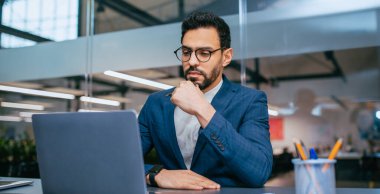 Mavi takım elbiseli bir adam bir masada oturup dizüstü bilgisayara bakıyor. Elinde bir kalem tutuyor ve elindeki göreve odaklanmış gibi görünüyor. Ofisin arka planında büyük pencereler ve yeşil bitkiler var..