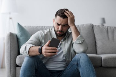 Psikolojik zorluklar, ilişki sorunları, depresyon ve kriz. Mutsuz, üzgün, sakallı, üzgün genç bir adam ellerini kaldırıp oturma odasında telefona bakıyor.