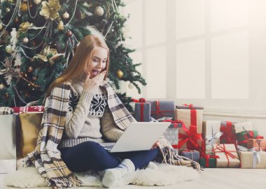 Dizüstü bilgisayarı olan sürpriz bir kız Noel ağacı arkasında oturuyor. Mutlu kadın, internetten kış indiriminde indirimli e-posta almış. Mevcut kutular arasında tatil evi olan, boşluk kopyala