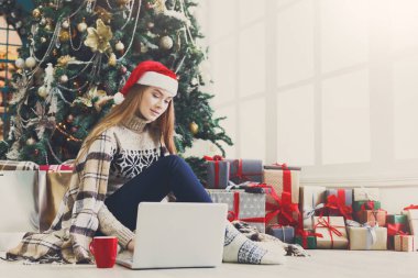 Kız ile dekore edilmiş Noel ağacı arka plan katta oturan laptop. Kahve içme ve tatil ev ortamında mevcut kutuları arasında online tebrik e-posta yazarken genç kadın. Kopya alanı