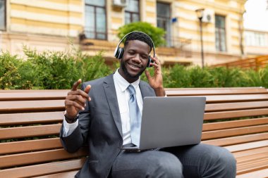 Heyecanlı Afro-Amerikan iş adamı, kablosuz kulaklıklar takıyor, modern laptop kullanıyor ve müzik dinliyor, ofis merkezinin yanındaki bankta oturuyor. Mutlu siyah menajer açık havada çalma listesini seviyor.