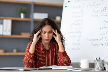 İşten yorgun düştüm. Baş ağrısı çeken, alnına ve şakaklarına dokunan, masa başında oturan, sorunları düşünen yorgun ve stresli genç bir kadının portresi.