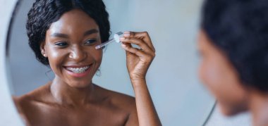 Bir kadın cildine serum uygularken bir damlatıcıyla gülümsüyor. Yuvarlak bir aynanın önünde ve iyi aydınlatılmış bir banyoda gibi görünüyor. Ortalık aydınlık ve temiz..