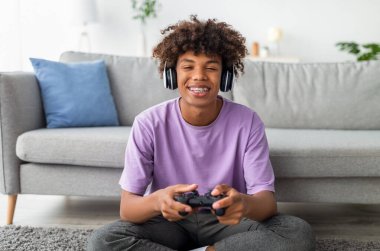 Evde kulaklık takan, çevrimiçi video oyunları oynayan havalı siyahi genç oyuncu portresi. Afrika kökenli Amerikalı ergenler kapalı alanda karantina sırasında eğlenceli bir hobi ediniyorlar.