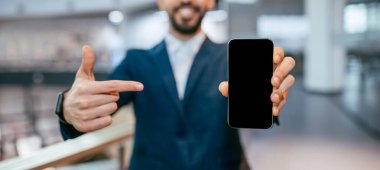 Bir adam iyi ışıklandırılmış bir binanın içinde, önünde siyah bir akıllı telefon tutuyor. Gülümser ve cihazı gösterir, bilgiyi ya da görüntüyü paylaşmaya hazırdır..