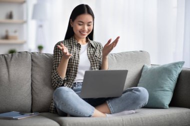 Güzel Asyalı bir bayan kanepeye oturmuş, neşeli bir şekilde el kol hareketi yapıyor, gülümsüyor COVID-19 kilitlenirken arkadaşlarıyla online parti yapıyor, mekanı kopyalıyor.