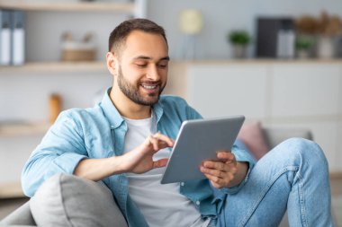 Tablet kullanan neşeli bir genç adam, evden internet üzerinden çalışıyor ya da çalışıyor. İş projesi yapan beyaz bir adamın portresi. Video görüşmesi ya da kapalı mekanda uzaktan görüşme.