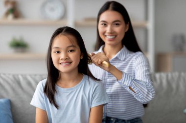 Asyalı bir annenin evdeki kanepede otururken saçlarını ahşap bir fırçayla taraması, kopyalama alanı, seçici odaklanma. Mutlu genç bir kadın çocuk saçıyla ilgileniyor.