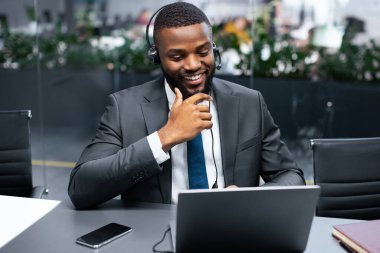 Masada takım elbiseli ve kulaklıklı bir adam oturuyor. Dizüstü bilgisayarına gülümseyerek bakıyor. Modern ofisin yeşil bitkileri ve gün ışığına çıkan büyük pencereleri var..