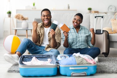 Tatil, sonunda. Neşeli Afrika kökenli Amerikalı çift, ellerinde seyahat biletleri ile evde bavullarıyla oturan kameraya gülümsüyor. Yeni Gezimizi Kutlayan Mutlu Turistler. Seçici Odaklanma