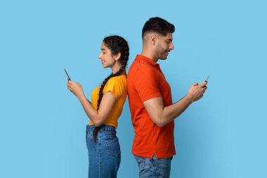 Sırt sırta verip akıllı telefonlarına odaklanmış genç bir adam ve kadın. Erkek turuncu bir gömlek giyerken kadın sarı bir bluz ve kot pantolon giyip rahat bir atmosfer yaratıyor..