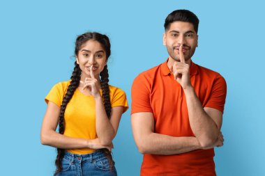 Genç bir adam ve bir kadın yan yana durur, her ikisi de gülümseyerek parmaklarını dudaklarına koyar sessizlik jesti ile..