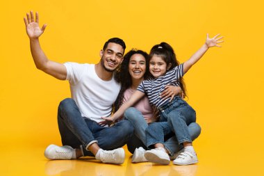 Sarı stüdyonun arka planında oturmuş eğlenen üç kişilik neşeli Arap ailesi, mutlu bir Orta Doğulu ebeveyn ve küçük kızları gülüyor ve ellerini kaldırıyor.