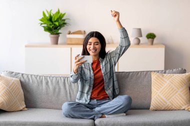 Genç Arap kadın bir koltukta oturuyor, akıllı telefonuna bakarken gülümsüyor. Heyecan içinde elini kaldırıyor, muhtemelen evinde neşeli bir ifadeyle zaferi kutluyor..