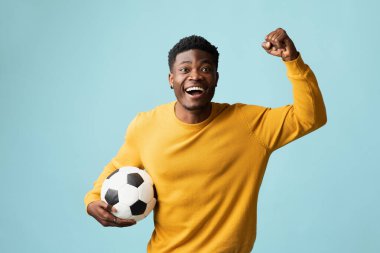 Duygusal milenyum siyahi bir adam futbol topuyla mavi stüdyo arka planında poz veriyor, Afro-Amerikan genç futbol fanatiği maça gidiyor, yumruğunu kaldırıyor, uzayı kopyalıyor. Spor konsepti