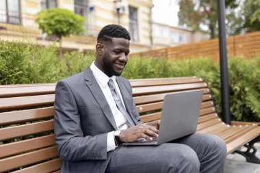 Uzaktan kumandalı iş. Kendine güvenen Afro-Amerikan iş adamı ya da takım elbiseli yönetici dışarıda ofis merkezinin yanındaki bankta otururken dizüstü bilgisayar kullanıyor.
